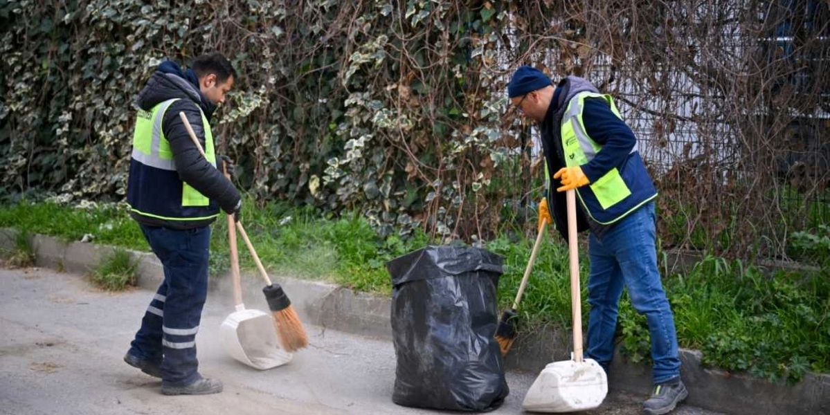 Üzerlerinde Bornova Belediyesi logolu fosforlu yelekler bulunan iki temizlik görevlisi, sokak kenarında süpürge ve faraş yardımıyla temizlik yapıyor. Görevlilerin arasında büyük, siyah bir çöp poşeti bulunuyor ve arka planda sarmaşıklarla kaplı bir çit yer alıyor.