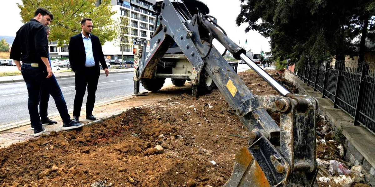 Siyah kıyafetli iki erkek, yol kenarındaki toprak alanda çalışma yapan bir iş makinesini inceliyor. İş makinesinin kepçesi toprağa doğru uzanmış durumda ve arka planda modern bir bina ile ağaçlar görünüyor.