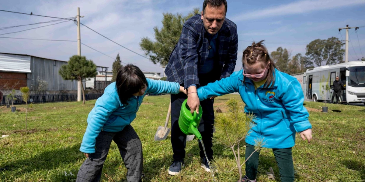 Mavi ceketli iki çocuk ve bir yetişkin, açık havada yeni dikilmiş bir fidanı yeşil bir sulama kabı yardımıyla suluyor. Çocuklardan biri sağ eliyle sulama kabının sapını tutarken, diğeri sol eliyle kabın ucunu yönlendiriyor; yetişkin ise ellerini çocukların ellerinin üzerine koyarak onlara destek oluyor. Arka planda çimenlik bir alan, ağaçlar ve park halindeki bir otobüs görülüyor.