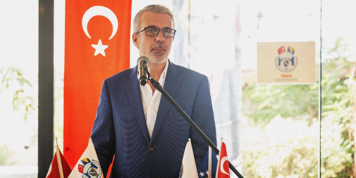 Lacivert ceketli, gözlüklü ve beyaz gömlekli bir iş insanı, önünde mikrofon bulunan bir kürsüde konuşma yapıyor. Arka planda dikey asılı bir Türk bayrağı ve cam üzerinde Türkiye ile Bosna Hersek bayraklarının yer aldığı bir logo bulunuyor. Masanın üzerinde ise çeşitli kurum bayrakları yer alıyor.