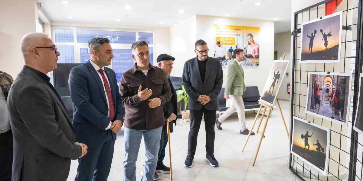 Aydınlık bir iç mekanda, bir grup erkek yan yana durarak metal bir pano üzerinde sergilenen Bergama temalı fotoğrafları inceliyor. Gruptakilerden biri el hareketleriyle açıklama yaparken, diğerleri dikkatle onu dinliyor. Panoda, kılıç ve kalkan kullanan figürlerin yer aldığı, Bergama'nın kültürel mirasını yansıtan fotoğraflar asılı duruyor. Arka planda ise başka bir kişi yürürken ve duvarda Bornova Belediyesi'ne ait bilgilendirici bir afiş görülüyor.