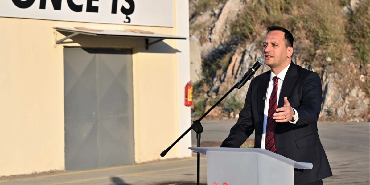 Koyu renk takım elbiseli ve çizgili kravatlı bir erkek, açık havada, üzerinde Bornova Belediyesi logosu bulunan bir kürsüde mikrofonla konuşma yapıyor. Arka planda üzerinde 