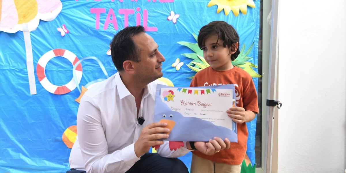 Beyaz gömlekli bir yetişkin, turuncu tişörtlü küçük bir çocuğa Bornova Belediyesi logolu bir katılım belgesi veriyor. Arka planda üzerinde 