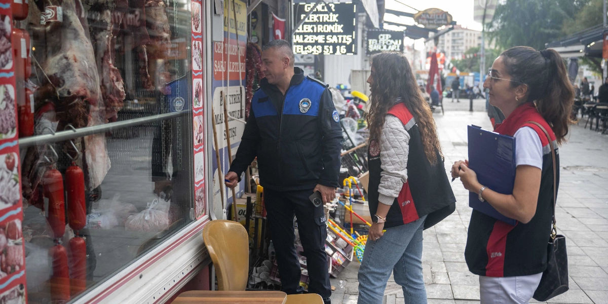 Bir zabıta memuru ve iki denetim görevlisi, bir kasap dükkanının önünde inceleme yapıyor. Zabıta memuru dükkanın vitrinine doğru bakarken, yanındaki iki kadın görevli ellerindeki dosyalarla not alıyor. Vitrinin arkasında asılı etler ve sucuklar görünüyor. Arka planda ise elektrik malzemeleri satan bir dükkan ve cadde üzerindeki diğer işletmeler yer alıyor.