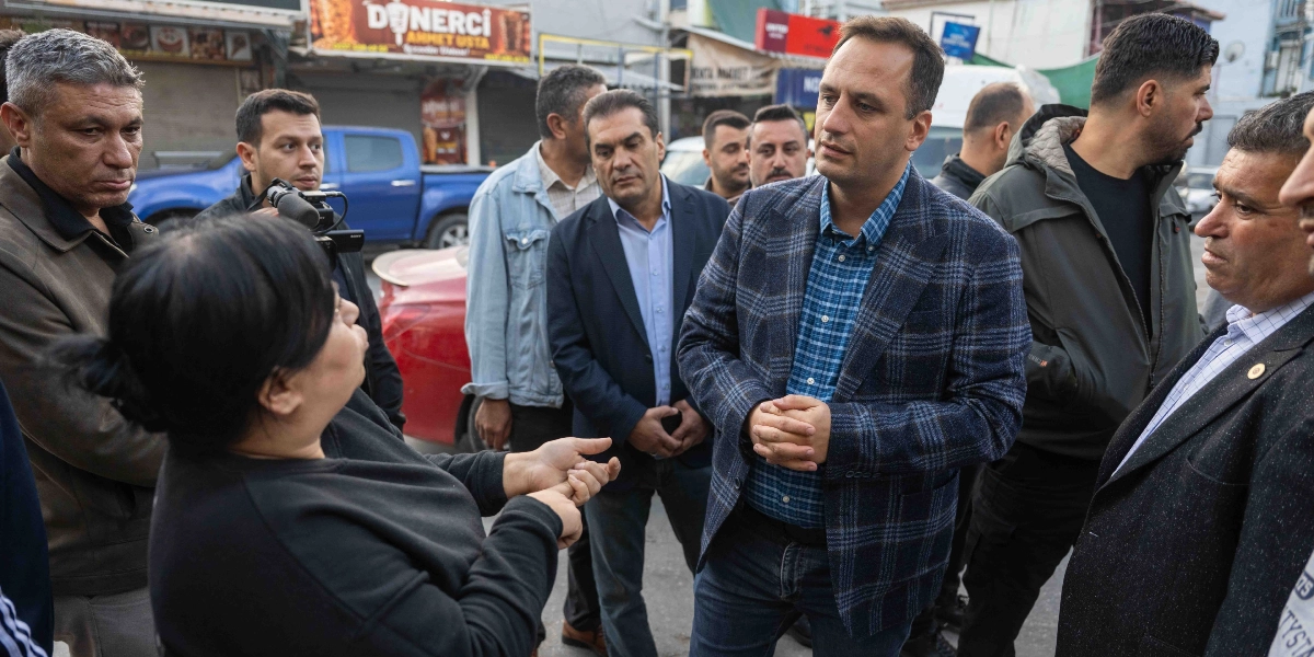 Bornova'da bir sokakta, ekoseli ceket giyen bir erkek, çevresindeki kalabalıkla birlikte bir kadını dinliyor. Kadın, elleriyle bir şeyler anlatırken, etraftaki diğer erkekler dikkatle onu izliyor. Arka planda dükkanlar, park halindeki araçlar ve 