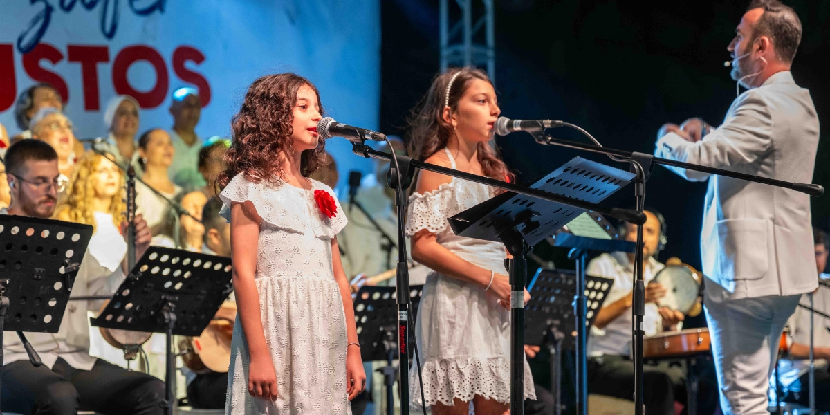 Açık havada düzenlenen bir konserde, beyaz elbiseli iki genç kız mikrofon başında şarkı söylüyor. Onların önünde notaların bulunduğu bir nota sehpası yer alırken, sağ tarafta beyaz takım elbiseli bir orkestra şefi orkestrayı yönetiyor. Arka planda ise müzisyenlerden ve koristlerden oluşan kalabalık bir grup bulunuyor. Sahne arkasındaki dev ekranda kırmızı harflerle yazılmış 