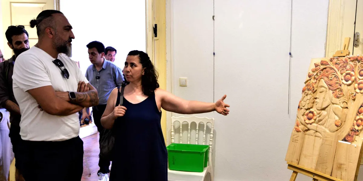 Bir sanat galerisinde, koyu renkli askılı elbise giymiş bir kadın, ahşap üzerine oyma tekniğiyle yapılmış, çiçekler ve insan figürü içeren bir eseri yanında duran sakallı bir adama eliyle işaret ederek anlatıyor. Arka planda ise iki erkek daha izleyici olarak yer alıyor.