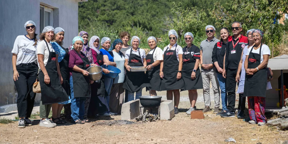 Açık havada, Bornova Belediyesi logolu siyah önlükler ve beyaz saç boneleri takmış bir grup kadın ve erkek, geleneksel yöntemlerle yemek hazırlığı yapıyor. Grubun ortasında, taşlar üzerine yerleştirilmiş bir ocak üzerinde büyük bir tencere bulunuyor. Bazı kadınlar ellerinde metal kaplar ve tepsiler tutarken, diğerleri yan yana dizilmiş şekilde gülümseyerek kameraya bakıyor. Arka planda ağaçlık bir alan ve beyaz bir yapı yer alıyor.