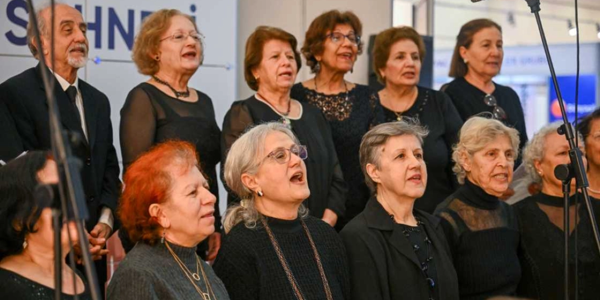 Siyah kıyafetler giymiş, çoğunluğu kadınlardan oluşan bir koro, önlerindeki mikrofonlar eşliğinde şarkı söylüyor. İleri yaş grubundaki koro üyeleri, odaklanmış bir şekilde performans sergiliyor.