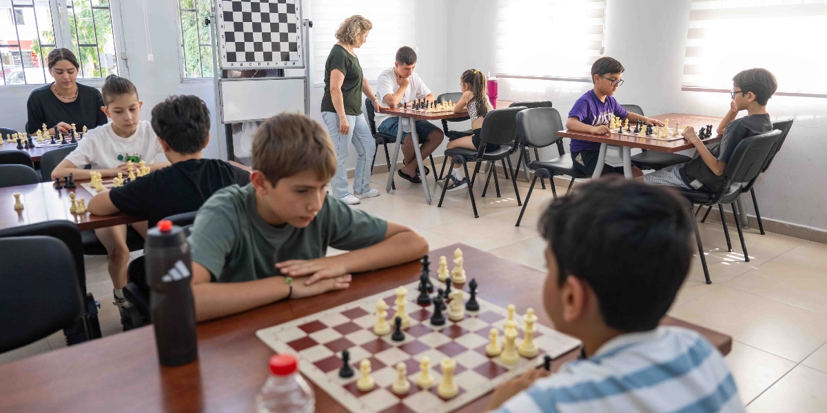 Aydınlık bir sınıfta satranç oynayan çocuk ve gençlerden oluşan bir grup. Masalara ikili gruplar halinde yerleşen öğrenciler, önlerindeki satranç tahtalarına odaklanmış durumda. Odanın arka kısmında ayakta duran bir eğitmen, öğrencileri gözlemliyor. Arka planda duvara asılı büyük bir gösterim satranç tahtası bulunuyor.