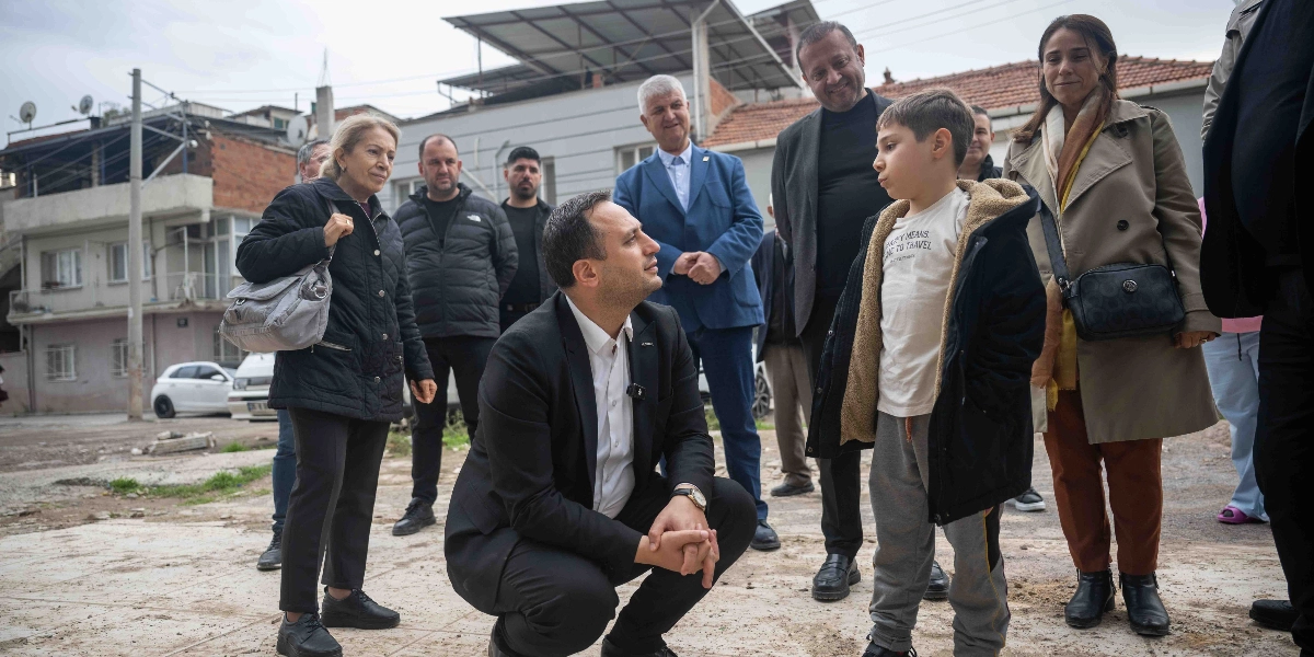 Bornova Belediye Başkanı Ömer Eşki, Çamdibi'nde bir mahalle ziyareti sırasında çömelerek küçük bir çocukla sohbet ediyor. Başkanın çevresinde belediye yetkilileri ve mahalle sakinleri yer alıyor. Arka planda ise yerleşim yerleri ve binalar bulunuyor.