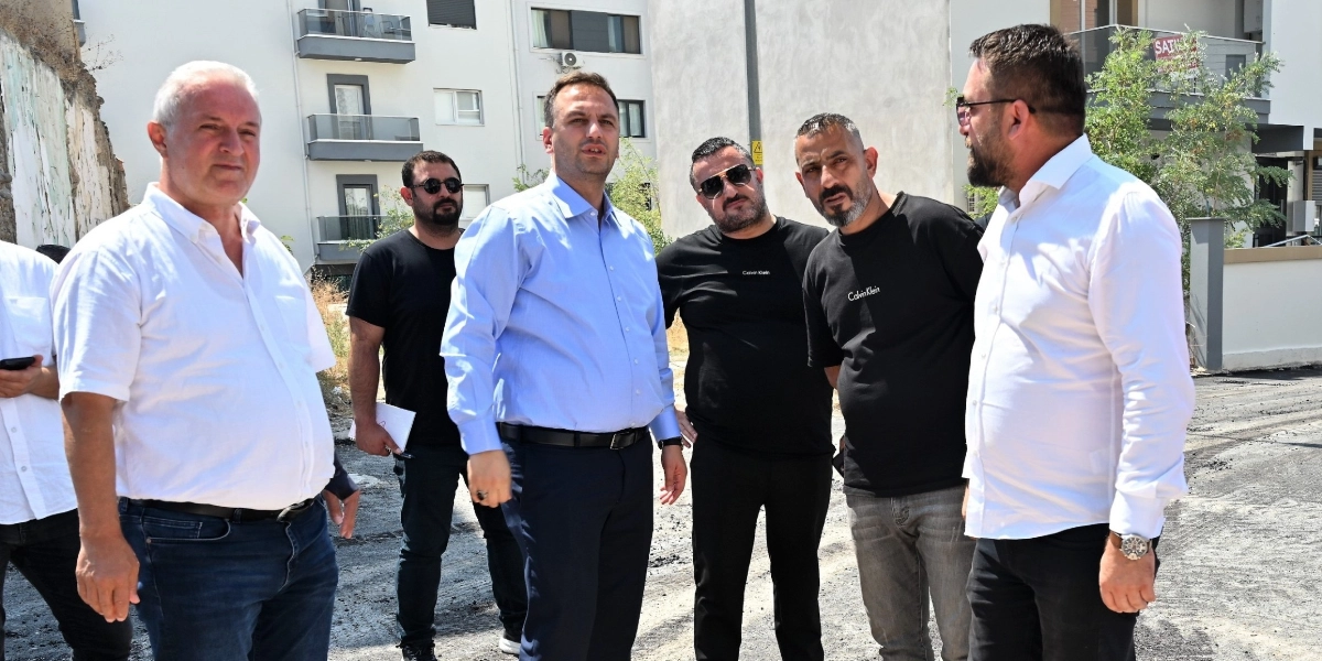 Açık havada, yeni yapılmış asfalt bir yolda yan yana duran ve bir projeyi inceleyen altı kişilik erkek grubu. Grubun ön planında açık mavi gömlekli bir yetkili, yanında beyaz gömlekli bir kişi ve siyah tişörtlü, güneş gözlüklü diğer katılımcılar yer alıyor. Arka planda ise modern apartman blokları ve çevre düzenlemesi yapılmış bir alan görülüyor.