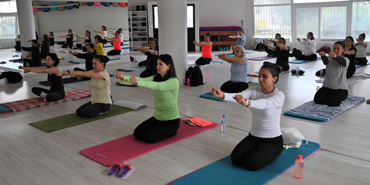 Geniş ve aydınlık bir spor salonunda, yoga matları üzerinde diz çökmüş şekilde oturan ve ellerini ileriye doğru uzatarak egzersiz yapan bir grup kadın. Kadınlar farklı renklerde spor kıyafetleri giymiş olup, salonun arka duvarındaki boydan boya aynada katılımcıların yansımaları görülmektedir.