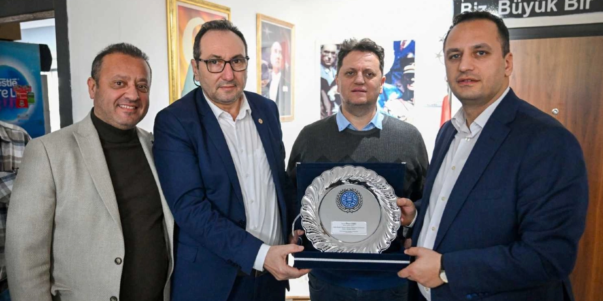 Dört erkek yan yana durarak, içlerinden biri tarafından tutulan gümüş renkli bir plaketi inceliyor. Soldan sağa doğru; bej ceketli, lacivert ceketli, gri kazaklı ve yine lacivert ceketli kişiler yer alıyor. Ortadaki iki kişi, üzerinde kurum logosu ve yazı bulunan gümüş plaketi birlikte tutuyor. Arka planda duvarda Atatürk portreleri ve 