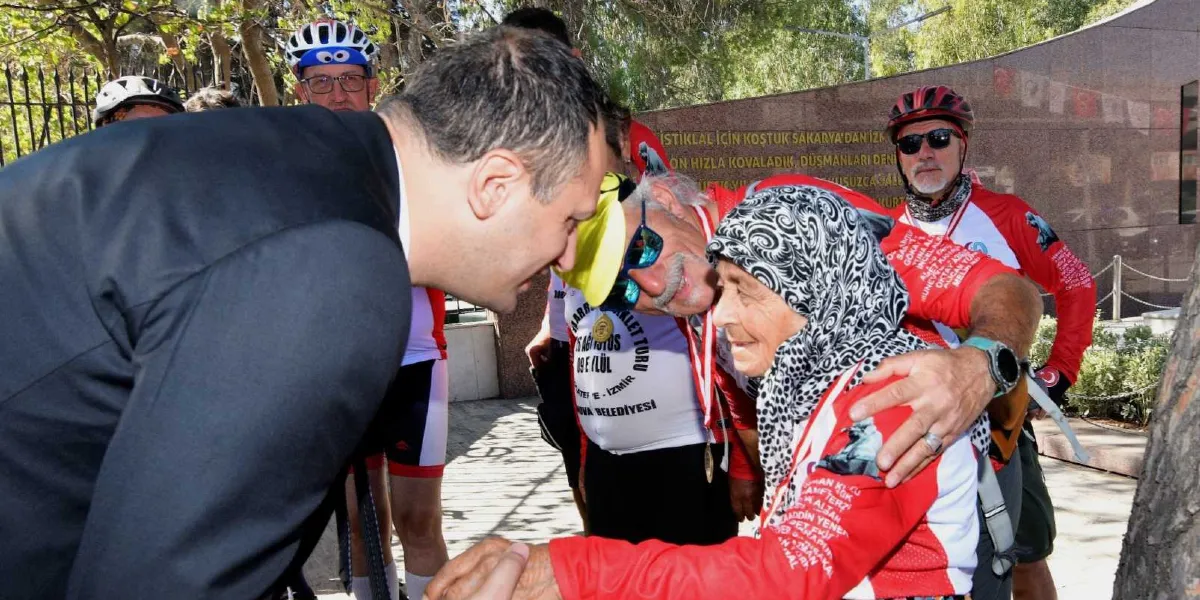 Siyah takım elbiseli bir yetkili, Bornova Belediyesi'nin düzenlediği 30 Ağustos - 9 Eylül Zafer Turu'na katılan, üzerinde özel tasarım kırmızı-beyaz formalar ve boyunlarında madalyalar bulunan bisikletçilerle bir araya geliyor. Yetkili, başörtülü yaşlı bir kadının elini tutarak onunla yakından ilgileniyor; yanındaki beyaz saçlı ve gözlüklü bir bisikletçi ise kadının omzuna elini koyarak ona destek veriyor. Arka planda, üzerinde 