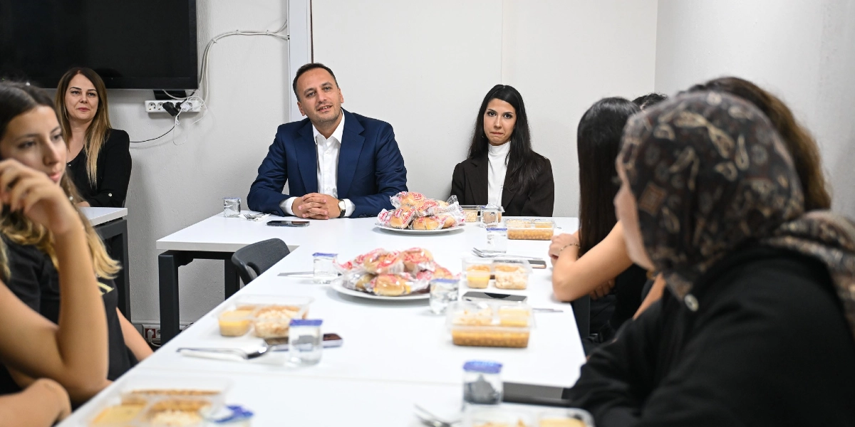 Lacivert ceketli bir erkek, beyaz bir masa etrafında toplanmış öğrencilerin arasında oturarak onlarla sohbet ediyor. Masanın üzerinde paketli yiyecekler ve plastik kaplarda yemekler bulunuyor. Öğrenciler ve diğer katılımcılar masanın farklı taraflarında oturmuş, dikkatle dinliyorlar.