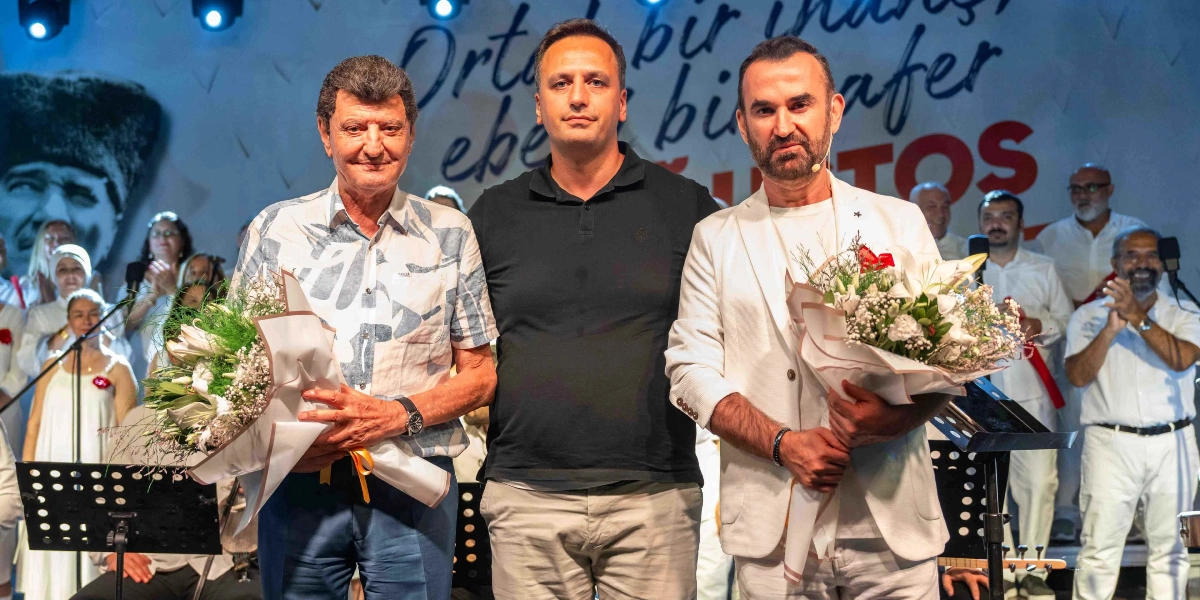 Sahne üzerinde beyaz kıyafetli bir koronun önünde duran üç erkek, ellerinde çiçek buketleriyle poz veriyor. Ortada siyah polo yaka tişörtlü bir adam, solunda desenli gömlekli, sağında ise beyaz ceketli bir adam bulunuyor. Arka planda 