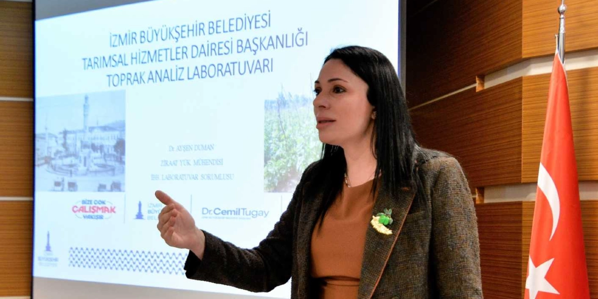 Ziraat Yüksek Mühendisi Dr. Ayşen Duman, İzmir Büyükşehir Belediyesi Tarımsal Hizmetler Dairesi Başkanlığı Toprak Analiz Laboratuvarı konulu bir sunum yaparken kürsüde konuşuyor. Arkasındaki yansıtıcı ekranda kurumun adı, kendi unvanı, İzmir Büyükşehir Belediyesi ve Dr. Cemil Tugay logoları ile 