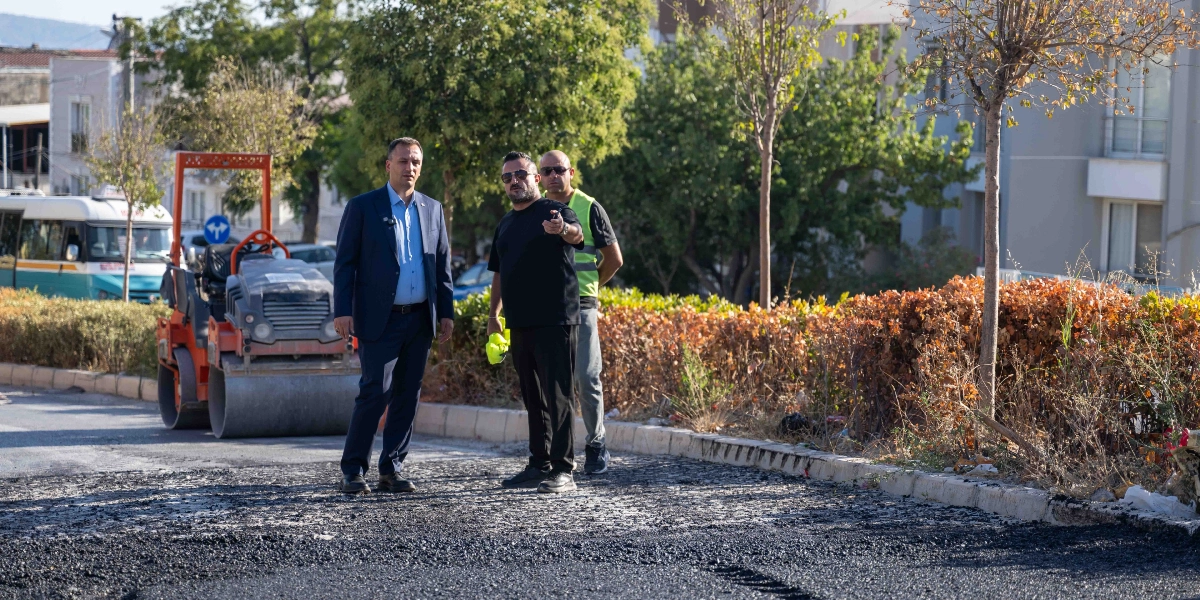 Bornova Belediyesi yetkilileri, asfalt çalışması yapılan bir yol üzerinde incelemelerde bulunuyor. Lacivert takım elbiseli bir yetkili, siyah tişörtlü ve güneş gözlüklü bir kişiyle yan yana dururken, arkalarında fosforlu yelek giyen bir görevli daha yer alıyor. Arka planda ise turuncu renkli bir silindir iş makinesi ve ağaçlıklı bir yol kenarı görülüyor.