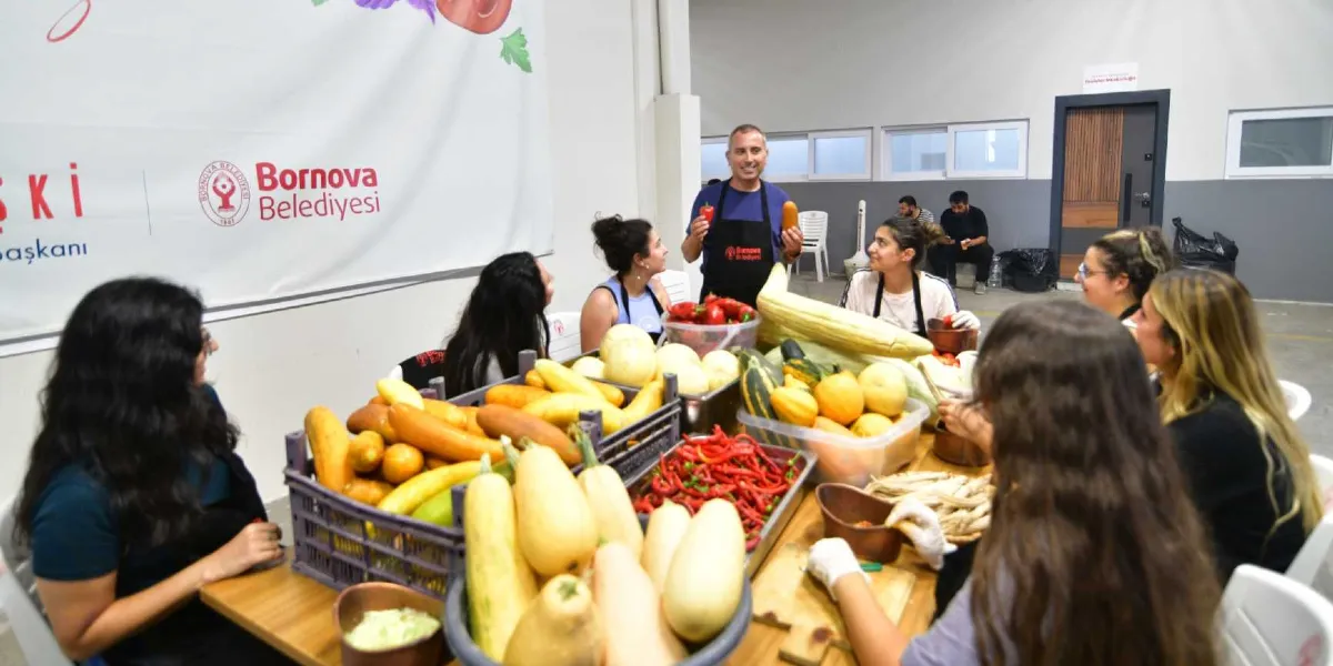 Bornova Belediyesi logolu bir pankartın önünde, üzerinde siyah önlük olan bir eğitmen, elinde tuttuğu sebzelerle etrafındaki katılımcılara atalık tohumlardan elde edilen ürünleri anlatıyor. Masanın üzerinde plastik kasalar içerisinde sarı kabaklar, kırmızı biberler ve çeşitli sebzeler bulunuyor. Katılımcılar masanın etrafında oturmuş, önlerindeki sebzeleri hazırlıyor veya eğitmeni dinliyor.