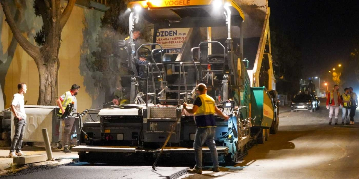 Gece saatlerinde bir cadde üzerinde asfalt serme çalışması yapan sarı renkli Vögele marka finişer iş makinesi ve çevresinde çalışan, reflektörlü yelek giymiş inşaat personeli. Arka planda bir kamyon, park halindeki araçlar ve yol kenarında duran insanlar görülüyor.