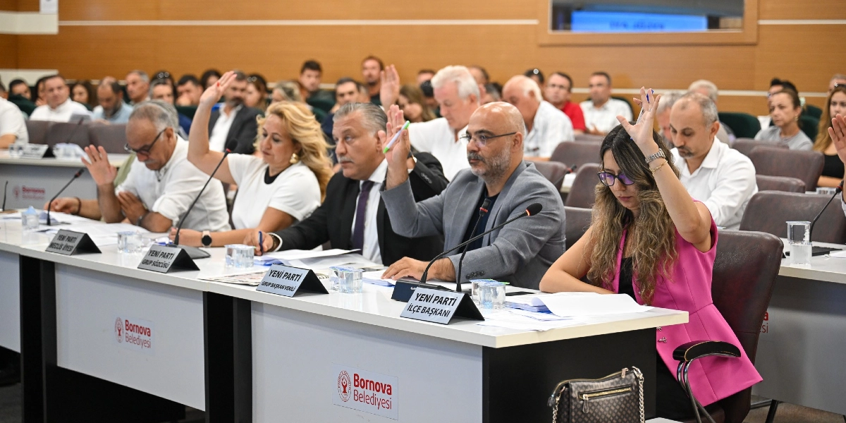 Bornova Belediyesi meclis salonunda düzenlenen bir toplantıda, masada oturan katılımcılar ellerini kaldırarak oylamaya katılıyor. Ön sırada, üzerinde 