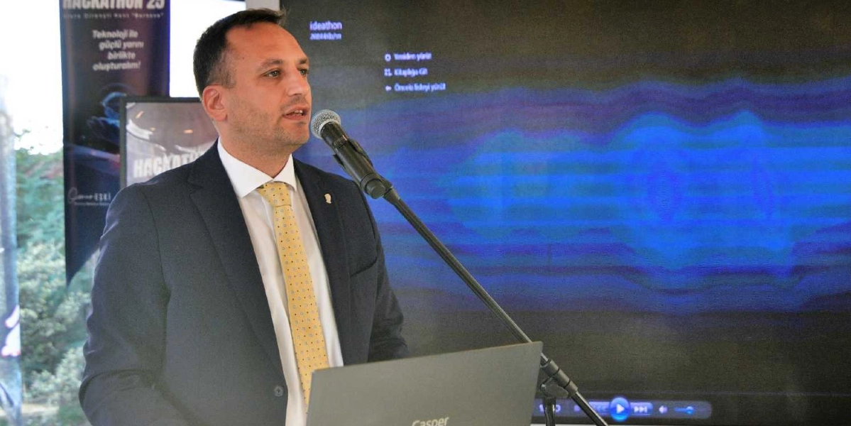 Koyu renk takım elbiseli ve sarı desenli kravatlı bir erkek, kürsüde mikrofon başında konuşma yapıyor. Önünde Casper marka bir dizüstü bilgisayar bulunuyor. Arkasındaki büyük ekranda 
