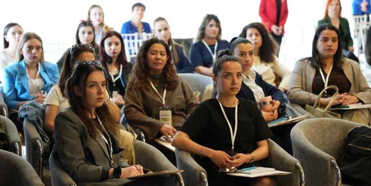 Modern bir konferans salonunda, boyunlarında yaka kartları bulunan ve dikkatle dinleyen, çoğunluğu kadınlardan oluşan bir grup katılımcı gri koltuklarda oturuyor. Ön planda, koyu renkli kıyafetler giymiş iki kadın, kucaklarında not defterleri ve kalemlerle sunumu takip ediyor. Arka sıralarda ise farklı renklerde kıyafetler giymiş diğer katılımcılar yer alıyor.