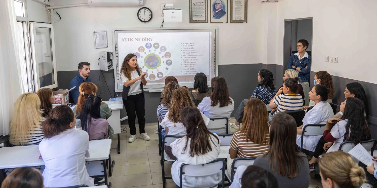 Bir sınıfta, akıllı tahtanın önünde duran bir eğitmen, atık yönetimi ve geri dönüşüm konulu bir sunum yapıyor. Tahtada 
