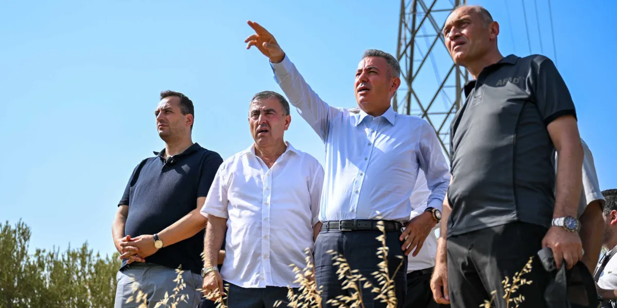 Açık havada, güneşli bir günde yan yana duran dört erkek, bir noktayı inceleyerek değerlendirmelerde bulunuyor. Açık mavi gömlekli bir kişi sağ eliyle ilerideki bir alanı işaret ederken, yanında beyaz gömlekli, lacivert tişörtlü ve üzerinde AFAD yazılı gri tişörtlü diğer kişiler dikkatle o yöne bakıyor. Arka planda yüksek bir elektrik direği ve mavi gökyüzü görülüyor.
