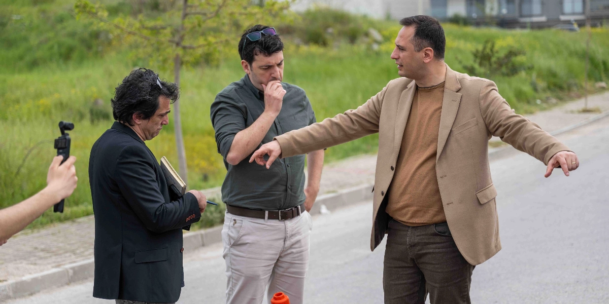 Açık havada, yeşil bir alanın yanındaki yolda duran üç erkekten biri, bej renkli ceketiyle eliyle yolu işaret ederek açıklama yapıyor. Yanındaki koyu yeşil gömlekli kişi onu dinlerken, siyah ceketli ve elinde not defteri tutan bir diğer kişi ise not alıyor. Sol tarafta, bir kişinin elinde kayıt cihazı tuttuğu görülüyor.