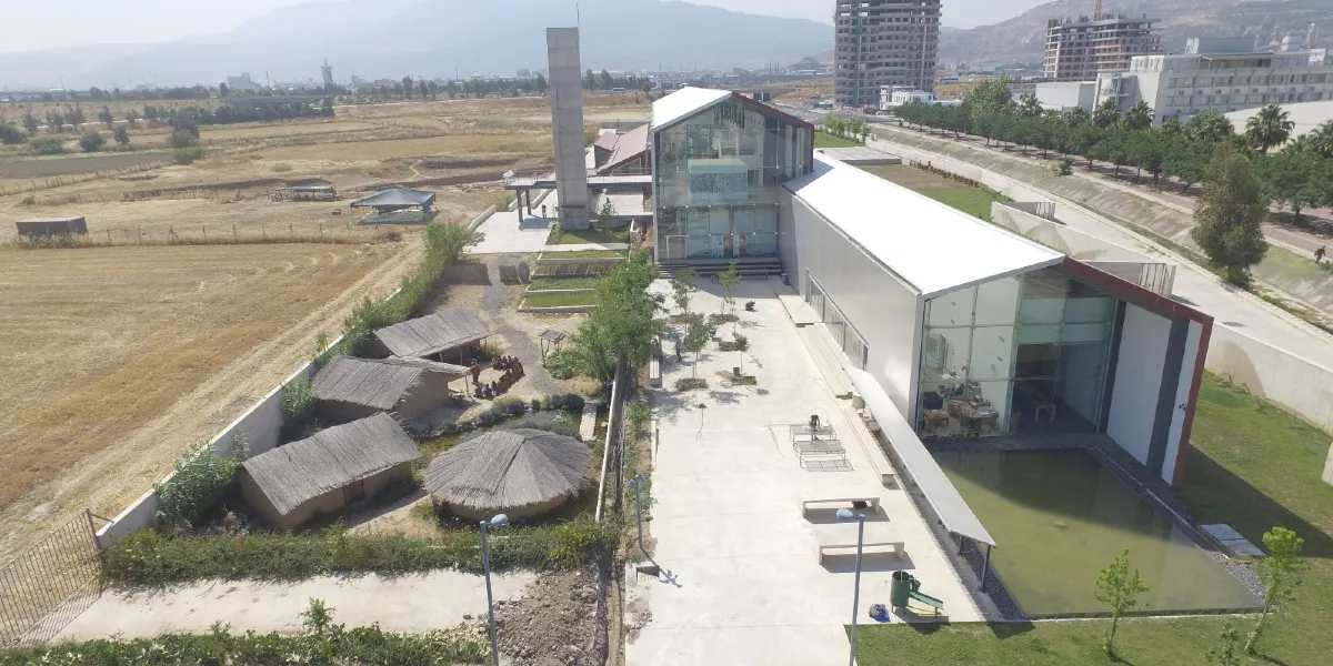 Yeşilova Höyüğü Ziyaretçi Merkezi'nin kuş bakışı görünümü; sağ tarafta modern mimariye sahip cam ve metal yapılı müze binası, önünde küçük bir süs havuzu ve oturma alanları yer alıyor. Sol tarafta ise tarih öncesi dönemi yansıtan saz çatılı geleneksel kulübeler ve açık hava sergileme alanı bulunuyor. Arka planda ise geniş bir arazi, ağaçlar ve uzakta şehir yapıları görülüyor.