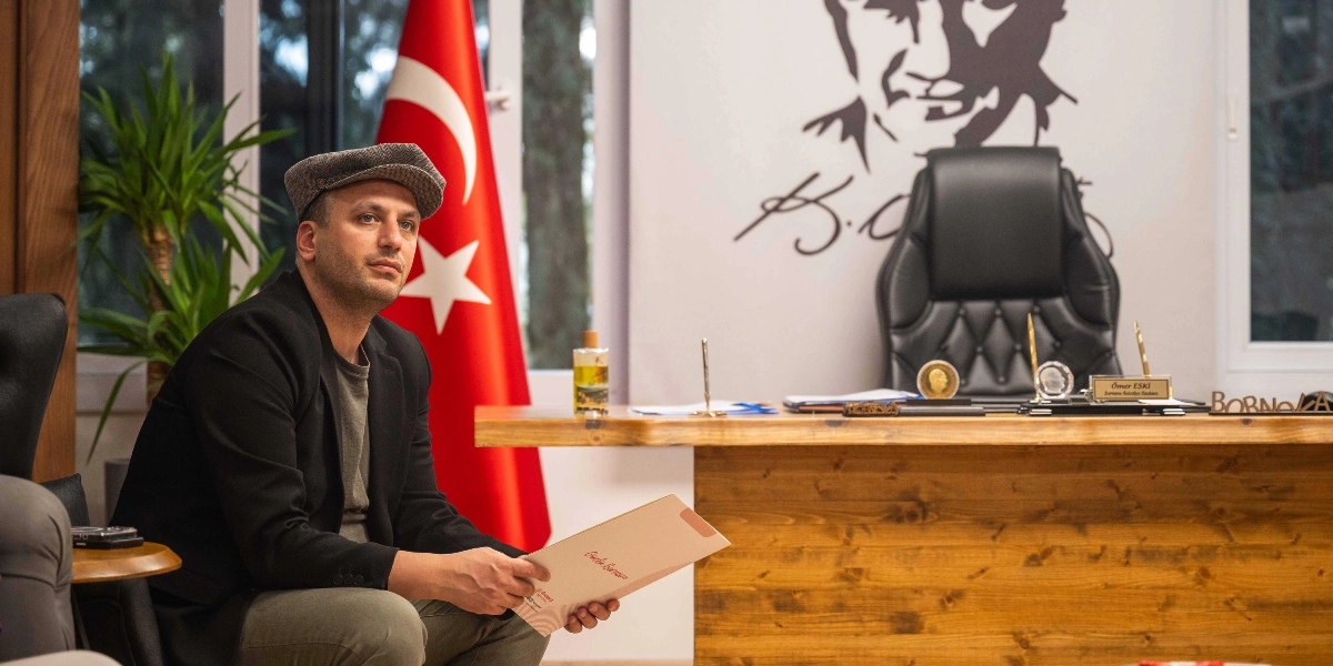 Kasket takan bir erkek, elinde bir dosya ile ofis ortamında oturuyor. Arka planda Türk bayrağı ve duvarda Mustafa Kemal Atatürk’ün silüeti yer alıyor. Ahşap çalışma masasının üzerinde Ömer Eski isminin yazılı olduğu bir isimlik ve çeşitli ofis aksesuarları bulunuyor.