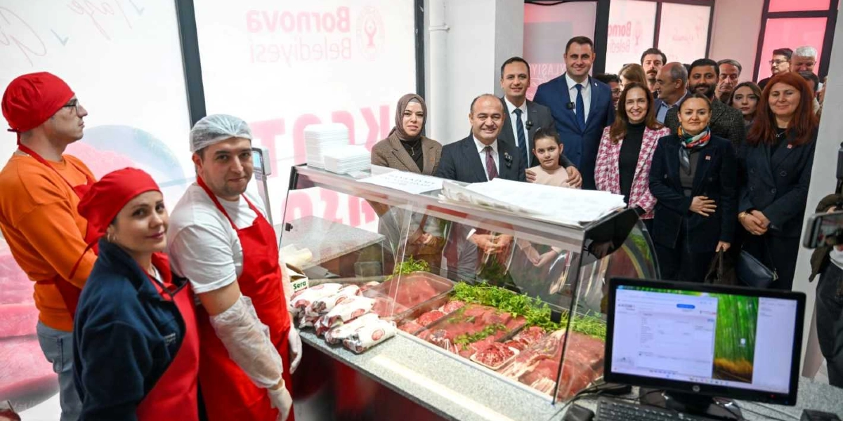 Bornova Belediyesi'ne ait bir kasap dükkanının açılışında, kırmızı önlük ve bone takan çalışanlar tezgahın arkasında dururken, yanlarında Bornova Belediye Başkanı ve beraberindeki heyet yer alıyor. Cam vitrinli tezgahın içinde taze et ürünleri ve yeşillikler sergileniyor, ön tarafta ise bir bilgisayar ekranı bulunuyor. Arka planda Bornova Belediyesi logosu ve yazıları görülüyor.