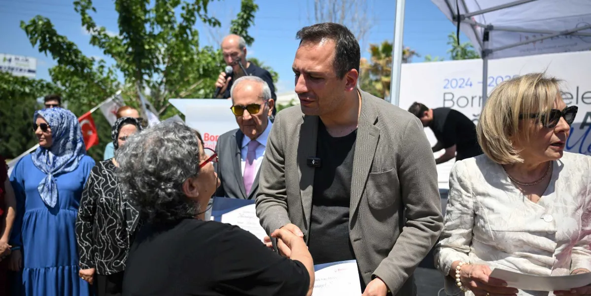 Bornova Belediye Başkanı Ömer Eşi, açık havada düzenlenen bir mezuniyet töreninde, hayat boyu öğrenme kurslarını başarıyla tamamlayan bir kursiyerle el sıkışarak sertifikasını veriyor. Arka planda diğer kursiyerler, protokol üyeleri ve üzerinde 