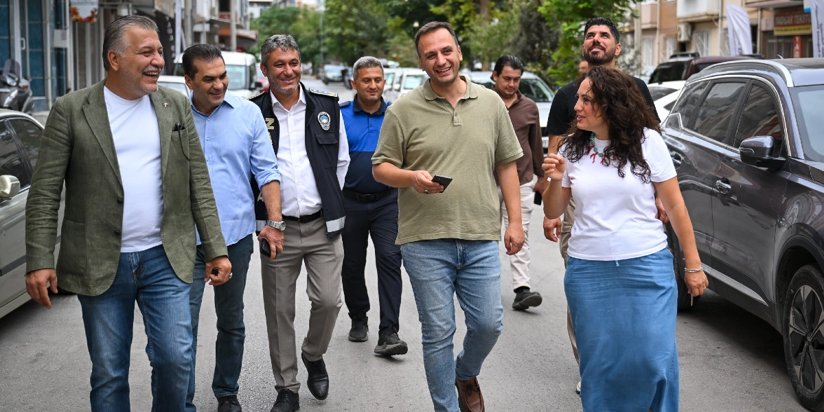 Bornova sokaklarında yürüyen ve sohbet eden, aralarında Bornova Belediye Başkanı Mustafa İduğ'un da bulunduğu bir grup yetkili ve vatandaş. Grup, park halindeki araçların bulunduğu bir caddede neşeli bir şekilde ilerliyor; önde yer alan kadın bir şeyler anlatırken, yanındaki yeşil tişörtlü kişi elinde telefonuyla onu dinliyor, diğer yetkililer ise arkalarında yürüyerek onlara eşlik ediyor.