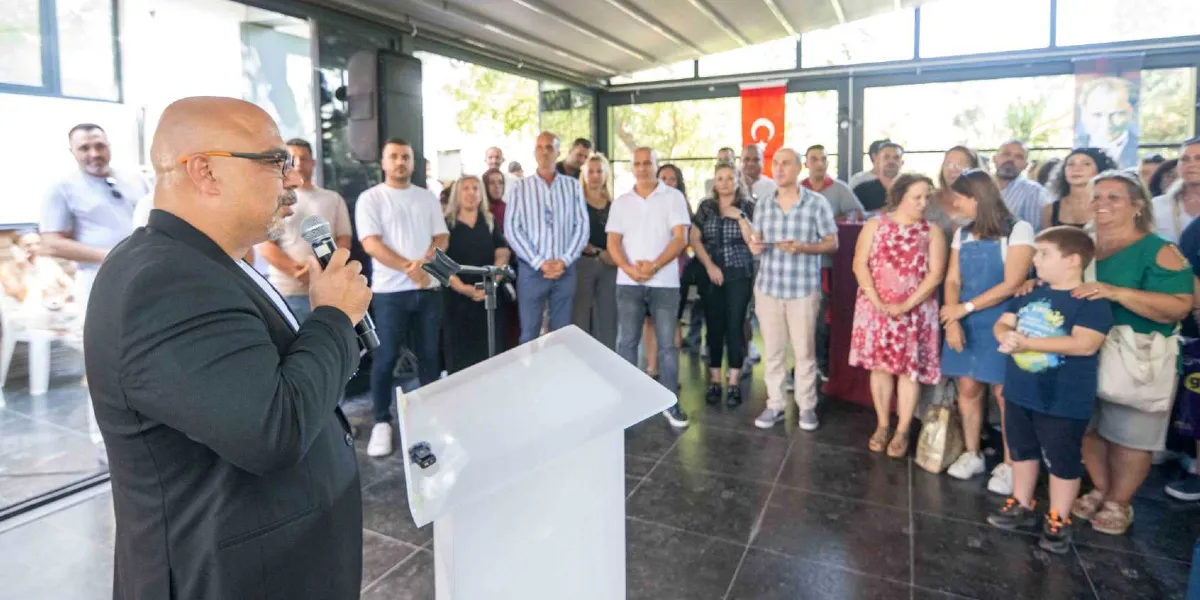 Kapalı bir mekanda, kürsü arkasında duran gözlüklü ve kel bir adam elinde mikrofonla konuşma yapıyor. Onu dinleyen, aralarında çocukların da bulunduğu kalabalık bir grup insan, kürsünün karşısında dikkatle duruyor. Arka planda Türk bayrağı ve Mustafa Kemal Atatürk'ün portresi yer alıyor.