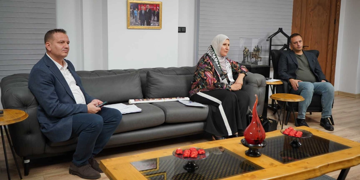 Modern bir ofis ortamında, gri bir koltukta oturan üç kişi bir toplantı halindedir. Sol tarafta lacivert ceketli bir erkek, ortada Filistin kefiyesi takan başörtülü bir kadın ve sağ taraftaki tekli koltukta ise koyu renk ceketli bir erkek yer almaktadır. Önlerinde ahşap bir orta sehpa, üzerinde kırmızı süs eşyaları ve dekoratif objeler bulunmaktadır. Arka plandaki duvarda çerçeveli bir fotoğraf asılıdır.