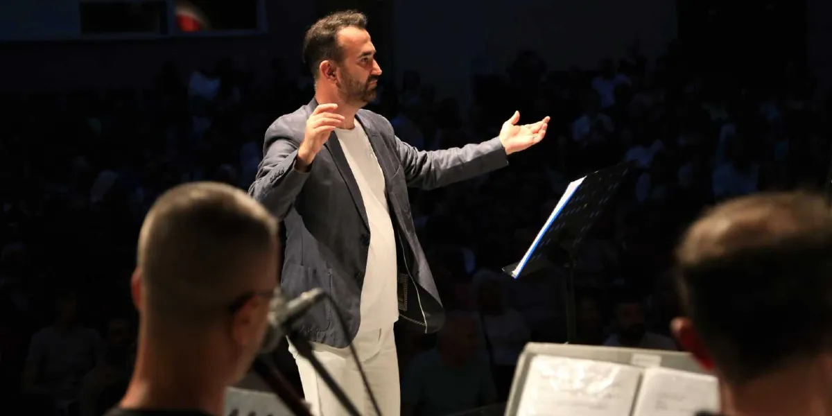 Koyu renkli bir ceket ve beyaz tişört giymiş bir orkestra şefi, sahne üzerinde kollarını iki yana açarak orkestrayı yönetiyor. Şefin önünde nota sehpası bulunurken, ön planda bulanık görünen müzisyenlerin sırtları ve arka planda ise karanlık bir ortamda izleyici kitlesi yer alıyor.