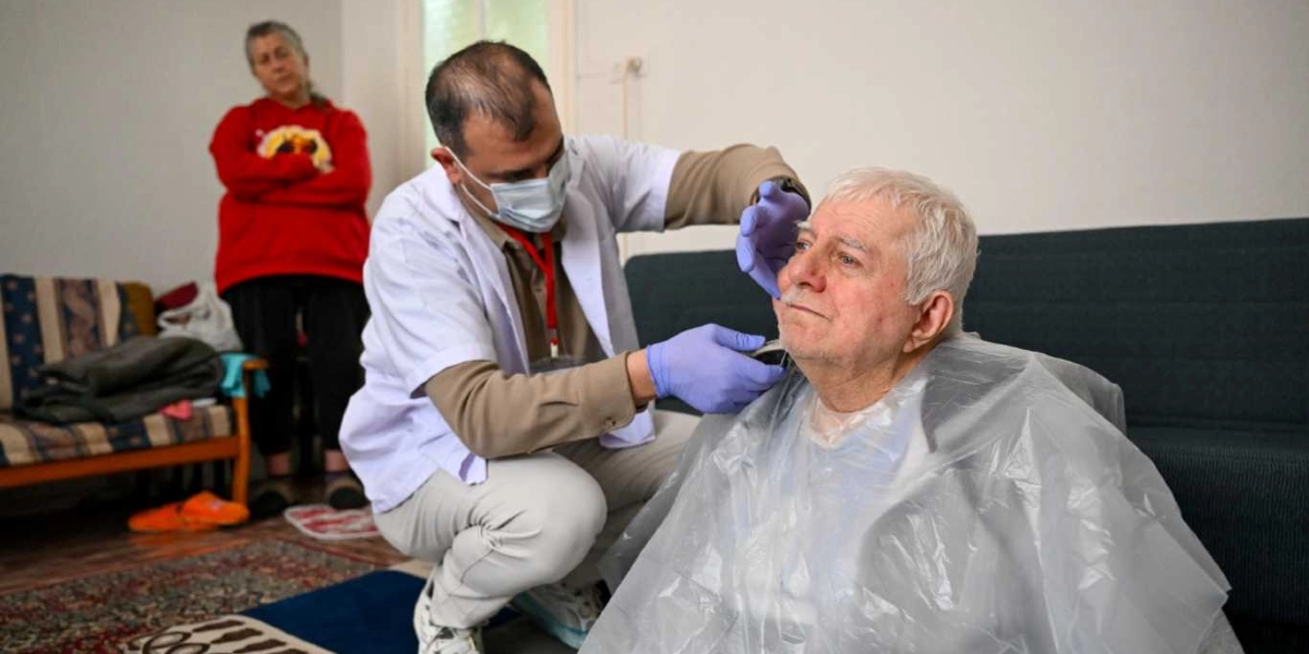 Beyaz önlüklü ve maskeli bir sağlık görevlisi, ev ortamında oturan yaşlı bir adamın yüzüne bakım yapıyor. Sağlık görevlisi mavi eldivenler takmış durumda ve yaşlı adamın üzerine koruyucu bir plastik örtü serilmiş. Arka planda kırmızı sweatshirt giymiş bir kadın ayakta durarak süreci izliyor.