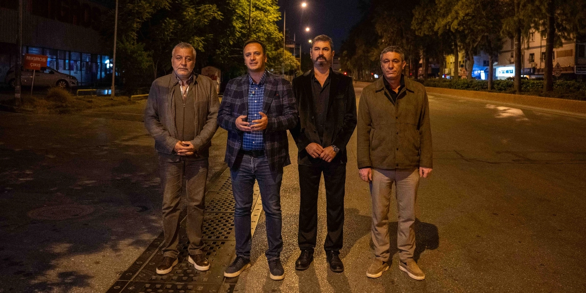 Gece vakti boş bir caddede yan yana duran dört erkek. Soldan sağa doğru; gri ceketli, kareli ceket ve mavi gömlekli, siyah ceketli ve bej ceketli dört kişi, Bornova'da kaldırımların düzenlenmesiyle ilgili bir çalışma kapsamında caddede poz veriyor. Arka planda ağaçlar, sokak lambaları ve bir marketin çıkış tabelası görülüyor.