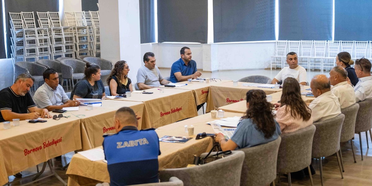 Bornova Belediyesi tarafından düzenlenen bir toplantıda, U harfi şeklinde birleştirilmiş masaların etrafında oturan muhtarlar ve belediye yetkilileri bir arada görülüyor. Masaların üzerinde Bornova Belediyesi yazılı örtüler serili. Ön planda sırtı dönük, üzerinde 