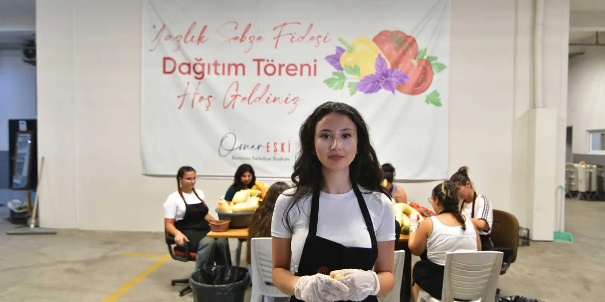Beyaz tişört ve siyah önlük giymiş, ellerinde şeffaf eldivenler bulunan genç bir kadın ön planda kameraya bakıyor. Arka planda, üzerinde 