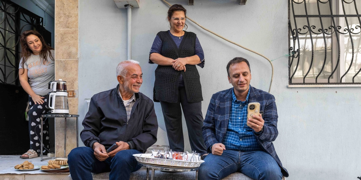Bir evin önünde oturan dört kişilik bir grup, ellerindeki akıllı telefona bakarak sohbet ediyor. Sağ tarafta oturan bir erkek elindeki telefonu gösterirken, yanındaki yaşlı adam ve arkasında ayakta duran kadın gülümseyerek ona eşlik ediyor. Soldaki kapı girişinde ise üzerinde 