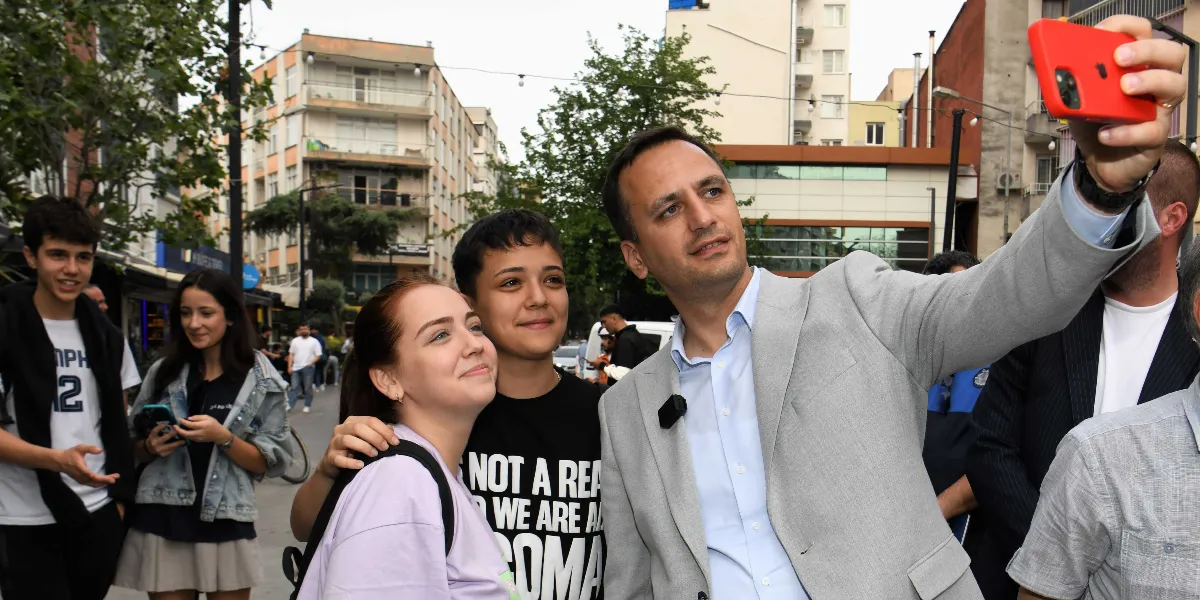 Gri ceketli bir erkek, dışarıda iki gençle birlikte kırmızı kılıflı bir telefonla selfie çekiyor. Arka planda Bornova sokaklarında yürüyen insanlar ve apartmanlar görünüyor.