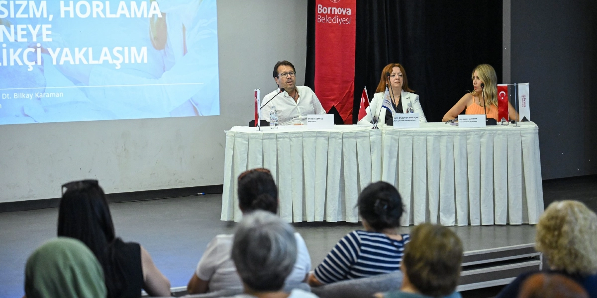 Bornova Belediyesi tarafından düzenlenen bir seminerde, beyaz örtülü bir masada oturan üç konuşmacı izleyicilere hitap ediyor. Soldan sağa doğru, beyaz gömlekli bir erkek, beyaz ceketli bir kadın ve turuncu bluzlu bir kadın yer alıyor. Masanın üzerinde mikrofonlar, küçük Türk bayrakları ve konuşmacıların isimlerinin yazılı olduğu kartlar bulunuyor. Arka planda yer alan dev ekranda 