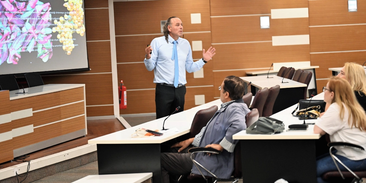 Modern bir konferans salonunda, açık mavi gömlekli ve kravatlı bir konuşmacı, yansıtılan bir sunum ekranının önünde durarak katılımcılara hitap ediyor. Konuşmacı ellerini kullanarak bir şeyler anlatırken, ön tarafta oturan üç katılımcı dikkatle onu dinliyor. Sunum ekranında mor sebzeler ve sarı üzüm salkımları gibi gıda görselleri ile besin değerlerine dair metinler yer alıyor.