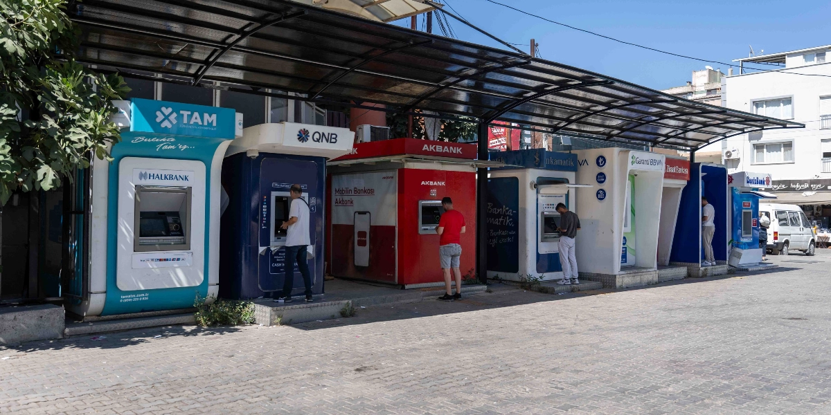 Bornova'da yan yana dizilmiş farklı bankalara ait ATM'lerin üzerini kapatan, güneşten ve yağmurdan koruyan geniş bir sundurma yapısı. Soldan sağa doğru Halkbank, QNB, Akbank, İş Bankası, Garanti BBVA, Ziraat Bankası ve DenizBank ATM'leri sıralanıyor; bazı ATM'lerin önünde işlem yapan kullanıcılar bulunuyor.