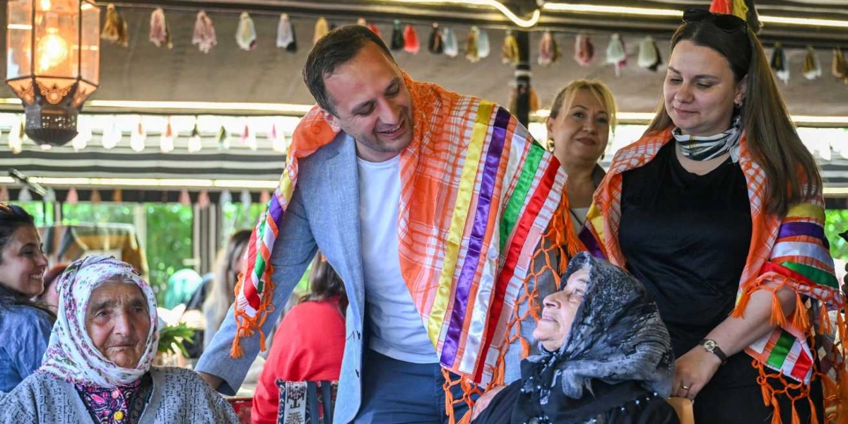 Bornova'da düzenlenen Anneler Günü etkinliğinde, omuzlarında renkli, püsküllü şallar bulunan bir erkek ve bir kadın, başörtülü yaşlı kadınlarla sohbet ediyor. Ortamda dekoratif fenerler ve süslemeler yer alıyor; katılımcılar samimi ve güler yüzlü bir ifadeyle birbirlerine bakıyor.