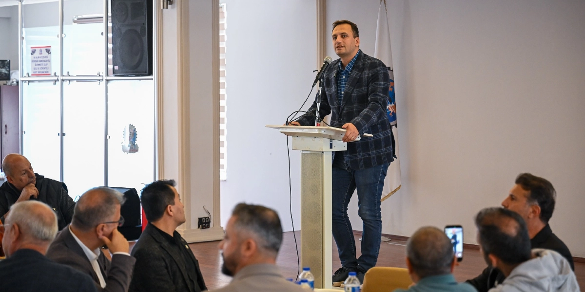 Kapalı bir salonda, kürsüde konuşma yapan ekose ceketli ve kot pantolonlu bir erkek, kendisini dinleyen bir grup katılımcıya hitap ediyor. Katılımcılar masalarda oturarak konuşmacıyı dikkatle takip ediyor, bazıları ise cep telefonlarıyla kayıt alıyor. Arka planda beyaz bir duvar ve üzerinde bir kurum logosu bulunan bayrak yer alıyor.